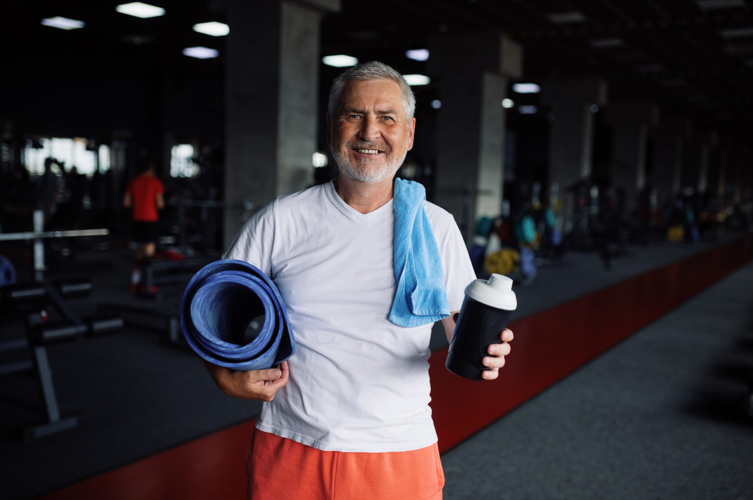 Old man with bottle of water, towel and mat in gym